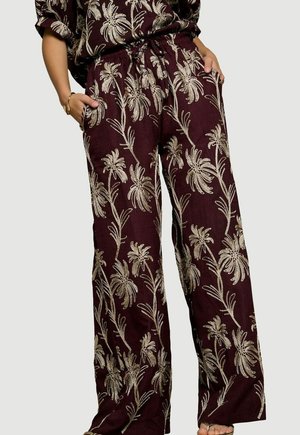 Pantalon large bordeaux avec broderie florale beige, porté avec un haut assorti, mains dans les poches et bracelet visible au poignet gauche.