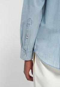 Camisa azul-claro com botões, feita de um tecido macio. Apresenta uma manga com punho pontiagudo e um botão, além de uma textura subtil, sendo rematada na parte inferior.