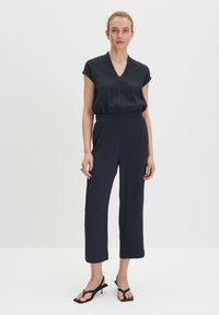 Marineblauwe jumpsuit met korte mouwen, V-hals en elastische tailleband. Voorzien van wijde pijpen en een gladde textuur. Gecombineerd met zwarte sandalen.