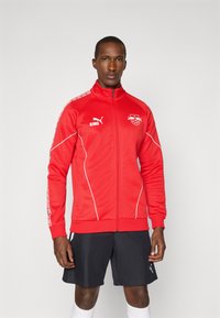 Red athletic zip-up jacket with white accents and Puma logo. Features high collar and patterned sleeves. Paired with black shorts and white socks.