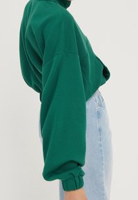 Zelená, př oversized mikina s vysokým límcem a nařasenými manžetami, doplněná o světle modré džíny s vysokým pasem. Měkká látka s matným povrchem.