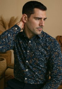 Donkerblauwe blouse met lange mouwen en een bloemenpatroon in bruine en witte tinten, voorzien van witte knopen en een standaard kraag.