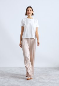 Λευκή κοντομάνικη μπλούζα με τυπωμένο λογότυπο, συνδυασμένη με ανοιχτό ροζ σατέν παντελόνια πυτζάμας που έχουν διακριτό καρό σχέδιο.