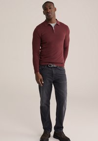 Dugih rukava bordo polo džemper s patentnim zatvaračem na ovratniku, u kombinaciji s tamnim trapericama i smeđim cipelama s vezicama. Glatka tekstura, ležerni kroj.