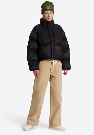 Kvinde iført sort dynejakke, beige vide bukser med snøre, hvide sneakers og olivengrøn hue med Vans-logo på hvid baggrund.