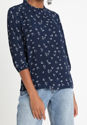 Blouse bleu marine à motif de feuilles blanches, col volanté, manches trois-quarts, associée à un jean denim bleu clair.