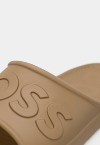 Sandale en caoutchouc couleur beige avec logo en relief, présentant une finition lisse, une semelle profilée et des bords arrondis. Semelle intérieure texturée pour une meilleure adhérence.