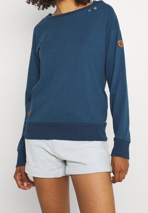 Kobieta w niebieskiej bluzie z długim rękawem, z guzikami przy kołnierzyku, oraz w jasnoszarych, podwiniętych spodenkach.