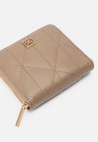 Μπεζ δερμάτινο πορτοφόλι με κουμπωτό σχέδιο, χρυσές λεπτομέρειες και κλείσιμο με φερμουάρ. Διαθέτει διαγώνια ραφή και μια μικρή λογότυπο λεπτομέρεια.