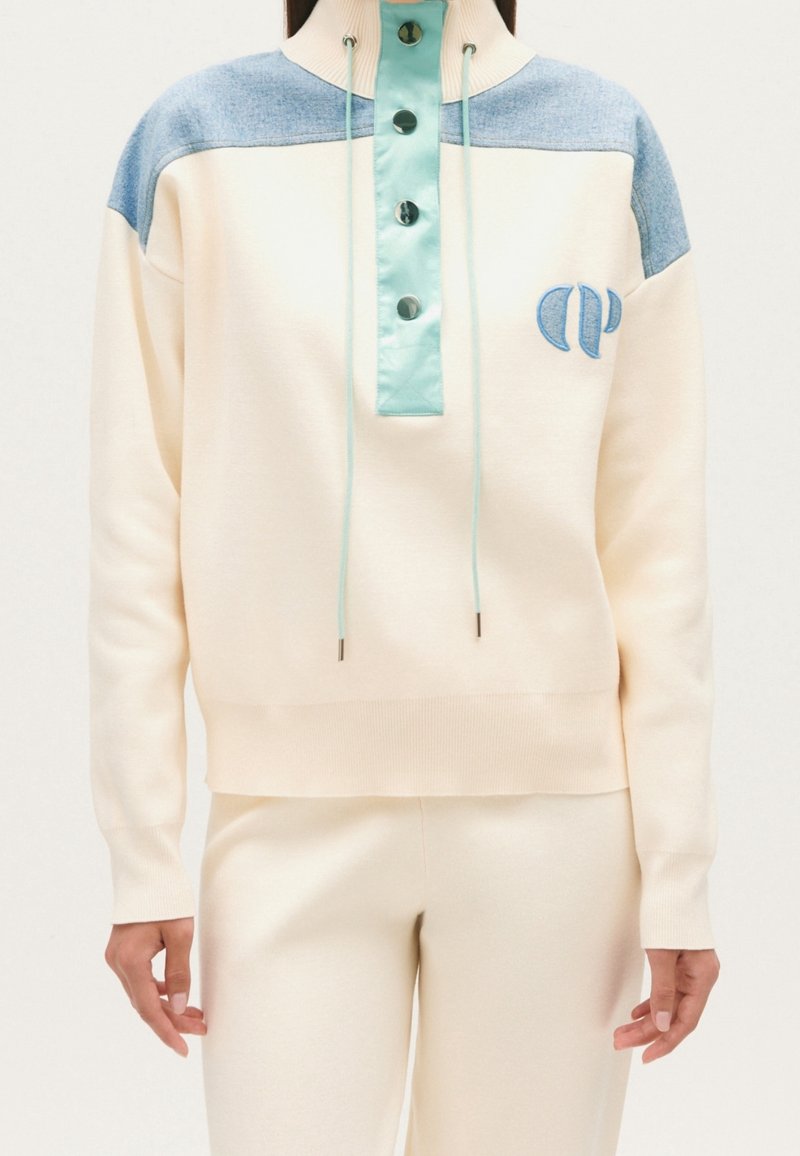 Femme portant un pull crème avec des panneaux bleu clair aux épaules, un col montant boutonné avec cordons, et un pantalon crème assorti.