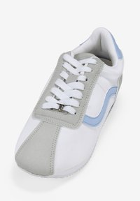 Zapatilla blanca con detalles en gamuza gris y acentos en azul claro, punta redonda, suela de goma y cordones planos con un diseño destacado en la lengüeta.