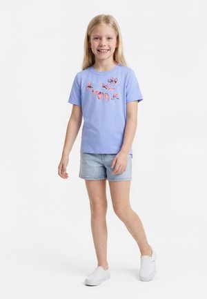 Νεαρή κοπέλα που χαμογελά, φοράει γαλάζια μπλούζα με λουλουδένιο λογότυπο, τζιν σορτς και λευκά παπούτσια slip-on, στέκεται μπροστά σε λευκό φόντο.