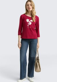 Rotes Langarmshirt mit weißen Blumenmotiven, kombiniert mit blauen Schlagjeans und weißen Turnschuhen, dabei eine beigetöne Tasche haltend.