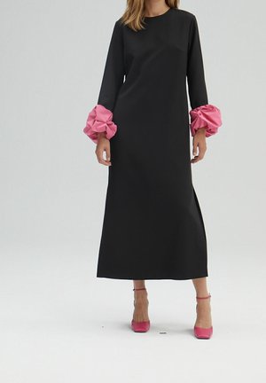 Frau trägt ein langes schwarzes Kleid mit pinkfarbenen, gerafften Manschetten und pinkfarbenen High Heels, steht vor einem schlichten weißen Hintergrund.