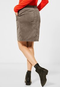 Jupe en velours côtelé gris avec poches arrière, longueur genou, associée à des bottines noires. La jupe présente une texture lisse et une taille ajustée.
