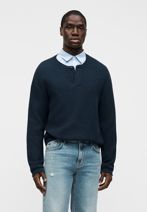 Marineblauer, gerippter Strickpullover mit Henley-Kragen, verfügt über eine einzelne Knopfleiste, getragen über einem hellblauen Hemd mit Kragen und kombiniert mit hellen Jeans.