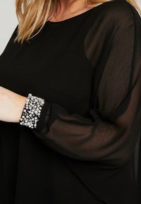 Haut noir avec des manches longues transparentes, présentant un poignet texturé orné de perles argentées et de strass. Tissu lisse avec une coupe fluide.