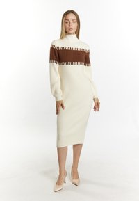 Strikket midi-kjole i creme og brun med ribbet struktur, der har en høj mock neck og lange ærmer. Neutrale spidse hæle fuldender looket.
