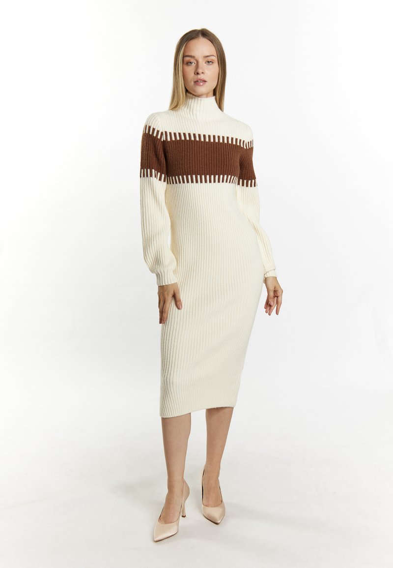Strikket midi-kjole i creme og brun med ribbet struktur, der har en høj mock neck og lange ærmer. Neutrale spidse hæle fuldender looket.
