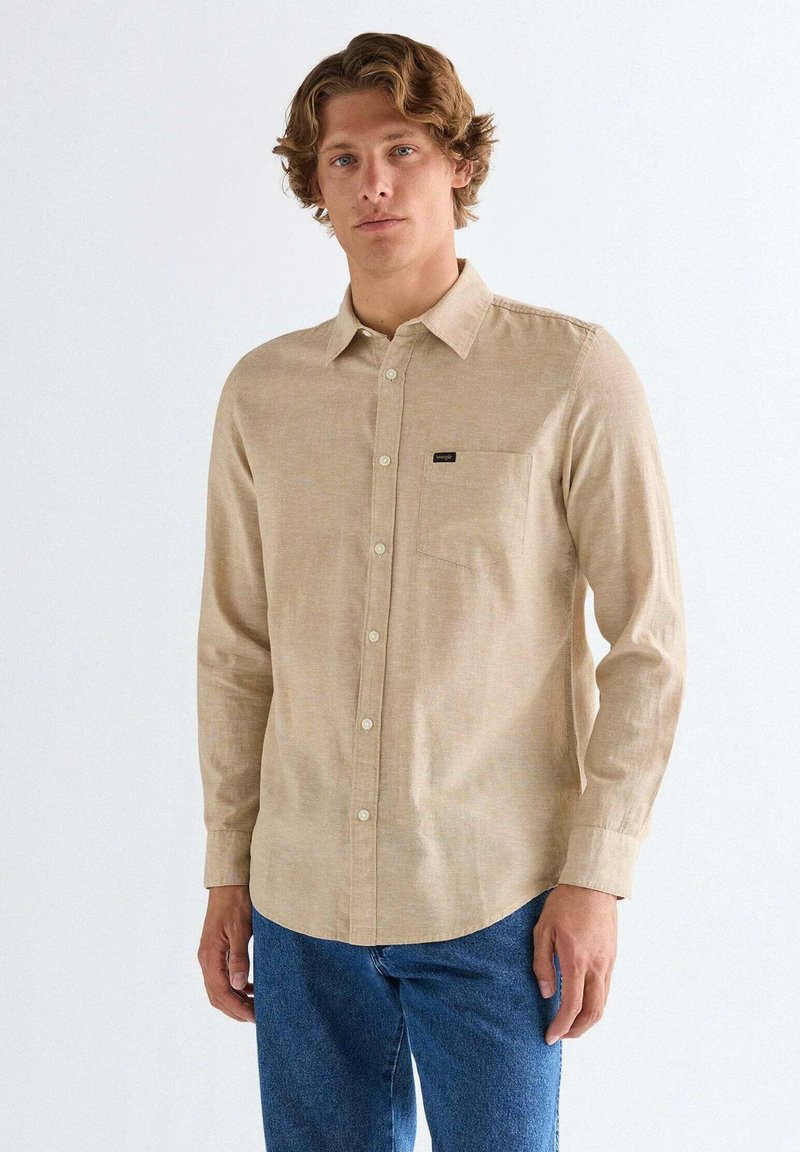 Jeune homme portant une chemise beige à manches longues boutonnée et un jean bleu, debout devant un fond blanc uni.