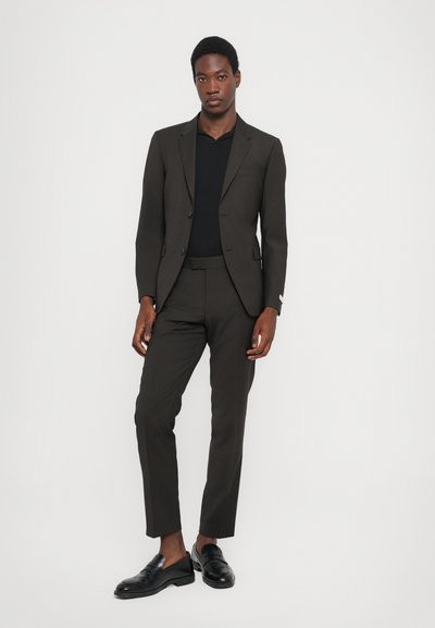 Tamno smeđi odijelo slim fit kroja, s jednorednim sakoom, sužavajućim hlačama i crnom košuljom. Crne mokasinke zaokružuju izgled.