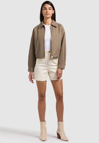 Femme aux cheveux longs et foncés portant une veste courte marron, un haut blanc, un short crème et des bottines beige, debout sur un fond neutre.