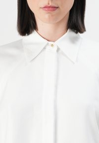 Žena s krátkými tmavými vlasy, oblečená v bílé texturované košili s špičatým límečkem a jedním zlatým knoflíkem na krku.
