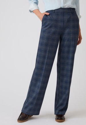 Vrouw draagt een hooggetailleerde blauw geruite broek met wijde pijpen, een lichtblauw overhemd en donkerbruine schoenen, staand met één hand in de zak.