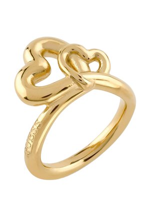 Pierścionek w kolorze złotym z dwoma połączonymi otwartymi sercami, gładką obrączką i wygrawerowanym napisem "GUESS" po boku.