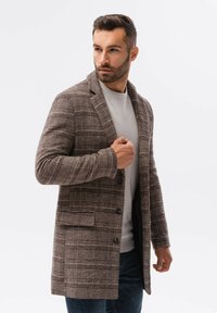 Ruskea ruudullinen takki tekstuurikankaasta, jossa on läppä, kaksi etutaskua ja kolme nappia, kerrostettuna vaaleanharmaan neuleen ja tummien farkkujen päälle.