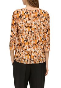 Vrouw in een losse, lange mouwtop met een abstract patroon in oranje, bruin en zwart en een klein sleutelgatdetail aan de achterkant.