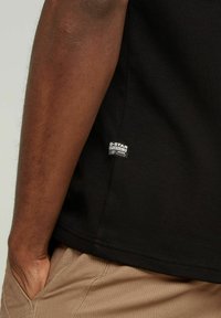 Camiseta de algodón negra con mangas cortas, que presenta una etiqueta de G-Star visible en el lado inferior. Con dobladillo plano, textura suave y ajuste casual.