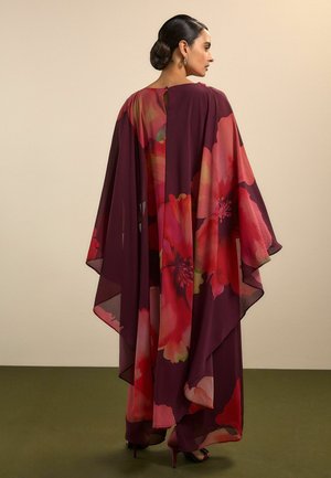 Žena so tmavými vlasmi v nízkom drdole má na sebe prúdiace šaty farby švestky s veľkými červenými a ružovými kvetinovými vzormi, stojaca bokom na olivovej podlahe.