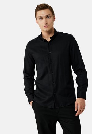 Jeune homme à la peau claire et aux cheveux courts bruns portant une chemise noire ajustée boutonnée à manches longues et un pantalon noir.