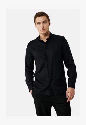 Junger Mann mit heller Haut und kurzen braunen Haaren, der ein eng anliegendes schwarzes Hemd mit langen Ärmeln und schwarze Hose trägt.