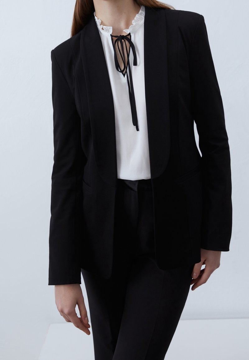 Femme portant un blazer noir et un pantalon avec un chemisier blanc à col volanté et un nœud noir noué au cou.
