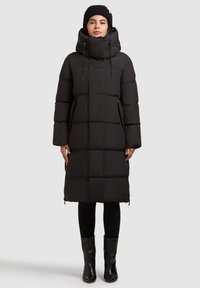 Sort lang pufferjakke med høj krave, quiltet design, snørehætte og sidelommer, parret med sorte tætsiddende leggings og ankelstøvler.