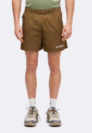 Homme portant un short marron Adidas Terrex, des chaussettes blanches et des chaussures de randonnée beiges, debout devant un fond uni.