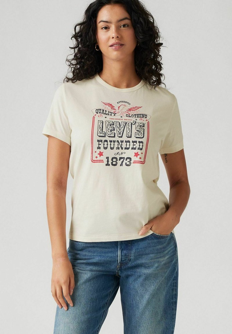 Vrouw met krullend haar, draagt een crèmekleurig Levi's t-shirt met rode en zwarte tekst en blauwe spijkerbroek, poseert met één hand in haar zak.