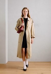 Beige trench coat dengan kancing besar, dipakai di atas gaun hitam. Model mengenakan kaus kaki putih dan sepatu hitam, sambil memegang tas tangan merah. Lantai kayu.