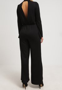 Mono negro con mangas largas, diseño de espalda abierta, cintura elástica y pantalones de pierna ancha. Confeccionado en tela suave. Tacones visibles.