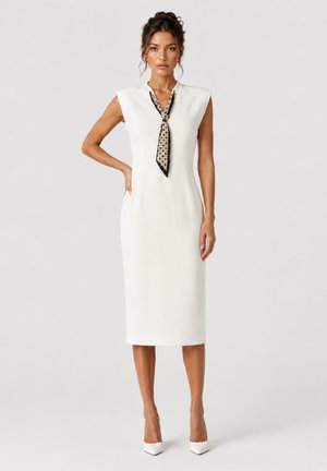 Woman wearing a sleeveless white midi dress with padded shoulders and a patterned necktie, standing with one hand on her hip.