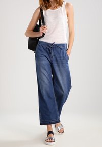 Débardeur blanc léger, pantalon en denim large bleu foncé à taille ajustable, et sandales multicolores. Sac à dos noir inclus.