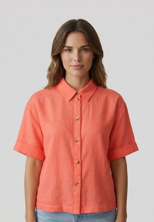 Vrouw met golvend bruin haar, gekleed in een koraalkleurig kort mouwen blouse met knopen en lichtblauwe spijkerbroek, staand voor een effen grijze achtergrond.