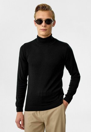 Zwarte coltrui gemaakt van zacht materiaal, met lange mouwen en een aansluitend design. Draagt zwarte ovale zonnebril voor contrast.