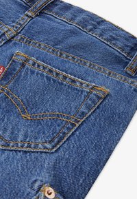 Ύφασμα τζιν μεσαίου μπλε τόνου, με πίσω τσέπη που έχει ραφές αντίθετου χρώματος και λεπτομέρεια με τρουκ, που αποτυπώνει κλασικό σχέδιο.