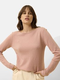 Różowy sweter z długim rękawem, wykonany z miękkiego, teksturowanego materiału, z okrągłym dekoltem, w połączeniu z jasnobeżowymi spodniami, ręce w kieszeniach.