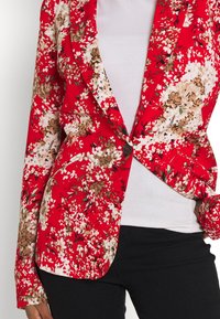 Červený květinový blazer s bílými a hnědými květy, s jedním knoflíkem, strukturovanými reverzy a dlouhými rukávy. Nosený přes bílé tričko.