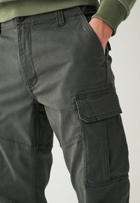 Dunkelgrüne Cargo-Hosen mit glatter Textur, ausgestattet mit mehreren Taschen und verstärkten Nähten. Der taillierte Schnitt sorgt für eine strukturierte Form.