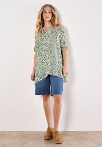 Camicia lunga verde e bianca a fantasia con scollo a V, abbinata a pantaloni corti in denim blu e zoccoli marroni, su uno sfondo neutro.